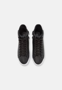 Paire de baskets montantes noires avec lacets noirs, fermetures éclairs latérales et semelles blanches, vues de dessus sur un fond uni clair.