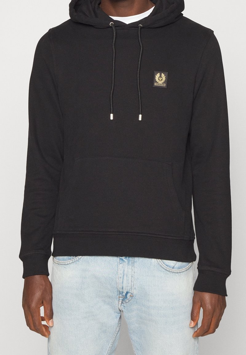 Personne portant un sweat à capuche noir avec une poche avant et un petit logo sur la poitrine, associé à un jean bleu clair.