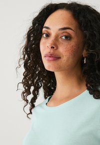 Lichtblauw t-shirt met een ronde hals, korte mouwen en zacht katoen. Het model heeft krullend haar en sproeten, met een zilveren ringoorbel.