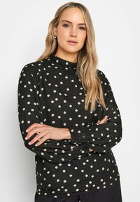 Långärmad svart blus med vita prickar, med hög halsringning och knäppte manschetter. Slät tygkvalitet med avslappnad passform.