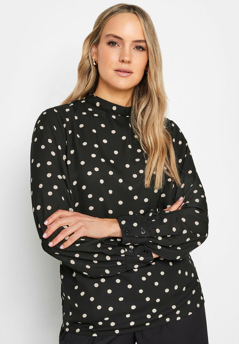Långärmad svart blus med vita prickar, med hög halsringning och knäppte manschetter. Slät tygkvalitet med avslappnad passform.