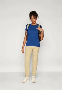 Blå kortermet t-skjorte, beige bukser med glidelås, og hvite joggesko. Ryggsekk med grå stropper over høyre skulder, stående foran en ensfarget bakgrunn.