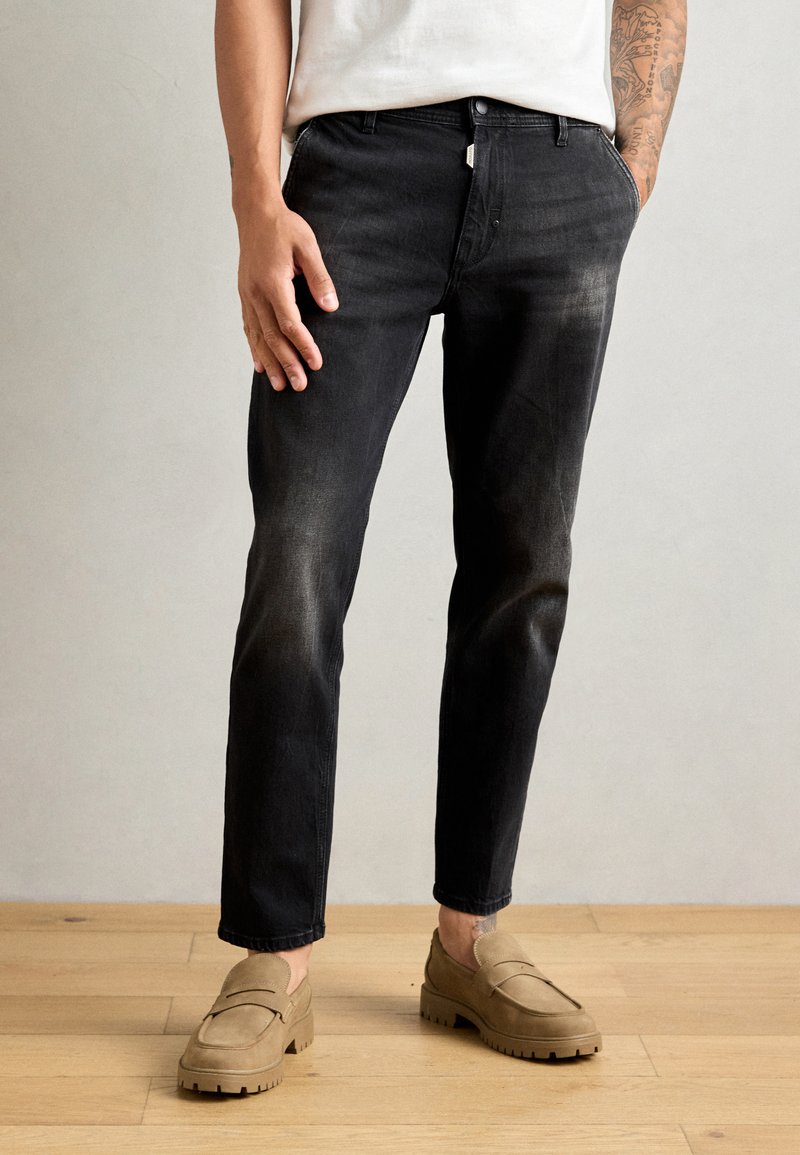 Jeans negri din denim cu croi slim, accente bleonate și bucle pentru curea, purtați cu pantofi slip-on bej pe un podea din lemn.