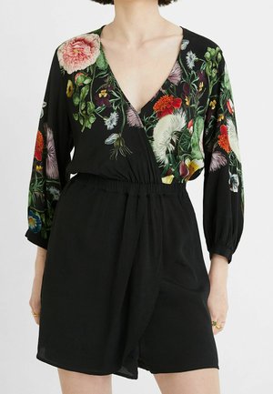 Nainen pukeutuneena mustaan romperiin, jossa on pitkät, kukkakuvioiset hihat ja V-pääntie. Yläosassa on eloisia, monivärisiä kukkia.