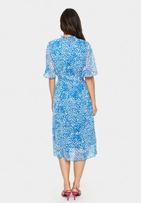 Robe à motifs bleus et blancs avec des manches courtes en tulle, un nœud à la taille et une jupe aux genoux. Associée à des sandales à talons hauts roses.