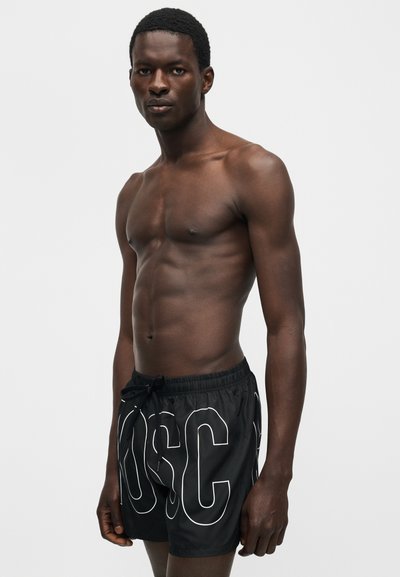 Αθλητικός άνδρας με σκούρο δέρμα που φοράει μαύρο μαγιό με μεγάλα άσπρα γράμματα περιγράμματος, στέκεται μπροστά σε ένα μονόχρωμο γκρίζο φόντο.