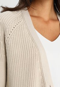 Béžový pletený cardigan s texturovaným vzorem, otevřeným předním dílem a výstřihem do V, s jemnými detaily na rukávech a okrajích.