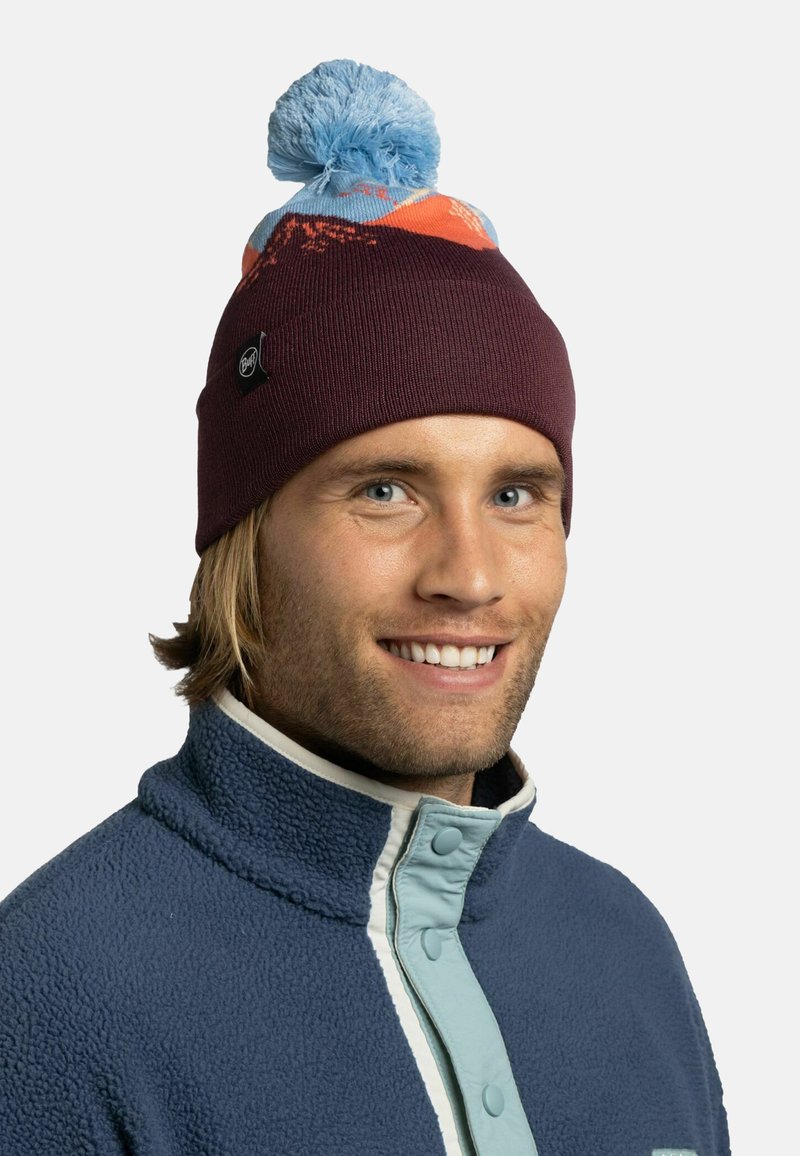 Gorro de punto burdeos con un pompón azul, que presenta un diseño de patrones en naranja y azul. El modelo lleva una chaqueta de forro polar con un cuello alto.