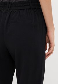 Pantalons noirs avec une taille élastique, en tissu doux et dotés d'une poche arrière, vus de côté avec une partie d'une main reposant dessus.