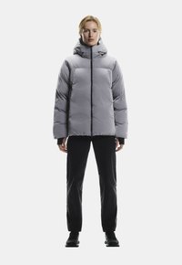 Szara kurtka puchowa z kapturem, z frontowym zamkiem błyskawicznym, elastycznymi mankietami i prostokątnym krojem, noszona na czarnych dopasowanych spodniach i butach.