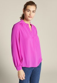 Rosa Langarmbluse mit eingeschnittenem V-Ausschnitt, gerafften Details an den Schultern und elastischen Bündchen. Leichter, glatter Stoff; lockere Passform.