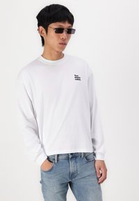 White long-sleeve t-shirt with a crew neck and black text "Too many rules." on the left chest, paired with light blue jeans.
