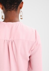 Ljusrosa blus med en plisserad baksida, mjuk textur och krage. Har stora guldiga hoopsörhängen.