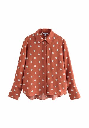 Chemise à manches longues orange rouille avec boutons, large col et pois blancs, ourlet légèrement incurvé, présentée sur fond blanc.