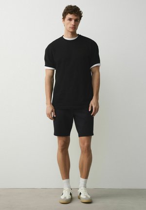 Young man standing against a plain wall wearing a black T-shirt with white trim, black shorts, white socks, and white sneakers.