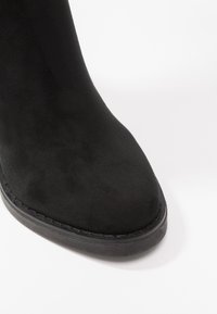 Stivale alla caviglia in suede nero con punta rotonda, texture liscia e suola in gomma. Dettaglio di cucitura delicata lungo il bordo della suola.