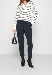 Gestreifter Pullover in Weiß und Marineblau mit V-Ausschnitt, kombiniert mit marineblauen maßgeschneiderten Hosen und schwarzen, hochhackigen Stiefeletten, während sie eine kleine schwarze Handtasche hält.
