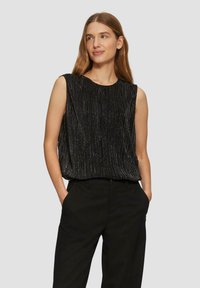 Haut sans manches noir avec des plis verticaux et des accents métalliques subtils. Le tissu a une texture douce et un design fluide. Associé à un pantalon sombre.