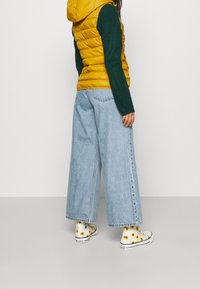 Gilet imbottito giallo, maniche in maglia verdi, ampi pantaloni in denim blu e sneakers bianche con motivo di girasoli. Vista posteriore dell'outfit.