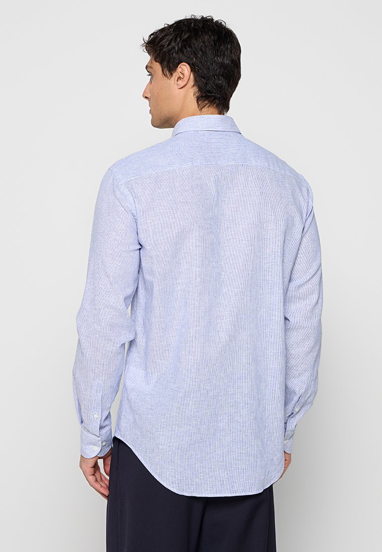 Hombre con cabello oscuro que lleva una camisa azul claro de manga larga abotonada visto desde atrás sobre un fondo liso.