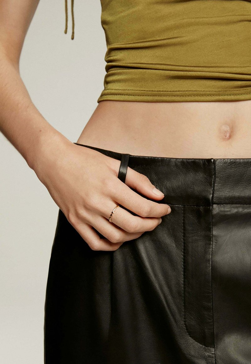 Falda de cuero negro con detalle de bolsillo lateral, combinada con una blusa ajustada de color verde oliva. Una mano con un anillo de oro reposa en la cintura.