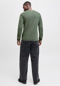 Man wearing an olive green long-sleeve sweater, loose black jeans, and black shoes, standing facing away against a plain light background.
