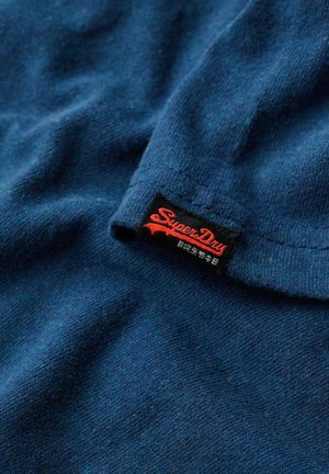 Mørkeblå tekstureret stof med en synlig sort etiket, der står "Superdry" i rødt. Materialet ser blødt og lidt meleret ud.