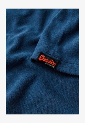 Mørkeblå tekstureret stof med en synlig sort etiket, der står "Superdry" i rødt. Materialet ser blødt og lidt meleret ud.