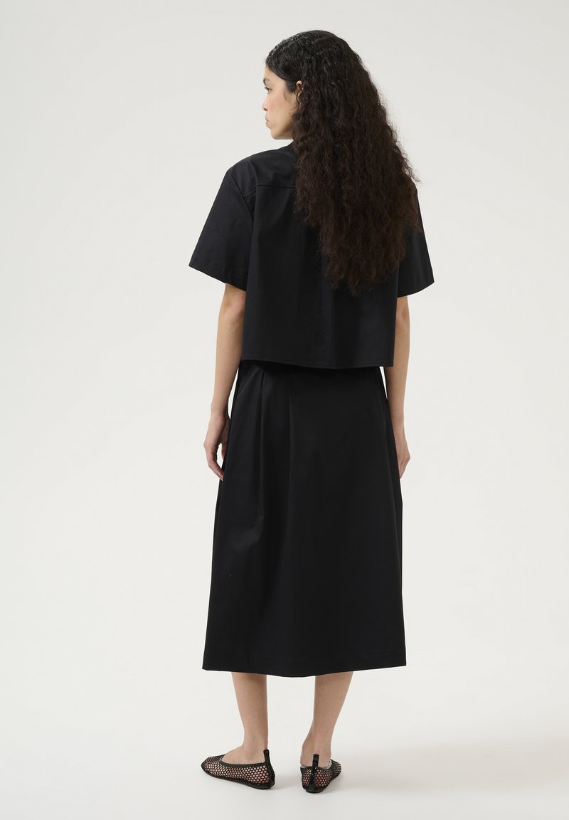 Top corto nero con maniche corte, abbinato a una lunga gonna nera. Indossato con ballerine nere in rete, che mostrano tessuto liscio e design semplice.