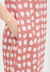 Un vestido rosa cuenta con grandes y irregulares lunares blancos, con una textura suave y bolsillos laterales integrados en el diseño.