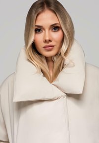 Světle béžový oversized kabát s velkým polstrovaným límcem, vyznačující se hladkou texturou a minimalistickým designem. Žádné viditelné kovové součásti nebo vzory.