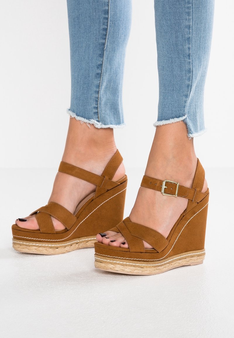 Pieds portant des sandales compensées en daim marron avec des brides à la cheville et un jean skinny bleu clair à ourlet effiloché sur un sol blanc.