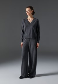 Temno siva pletena jopica z V-izrezom in rebrastimi manšetami, v kombinaciji s širokimi hlačami iz istega materiala, oboji narejeni iz mehkega blaga.