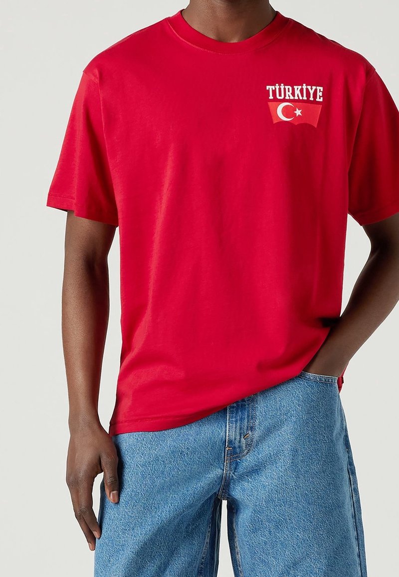 Homme portant une chemise rouge à manches courtes avec "Türkiye" et un graphique de drapeau sur la poitrine, associée à un jean en denim bleu clair.