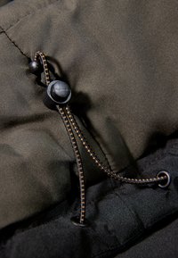 Svart jackdetalj med ett justerbart togglesystem med en texturerad snodd, framhävd av orange sömmar mot en slät tygbakgrund.