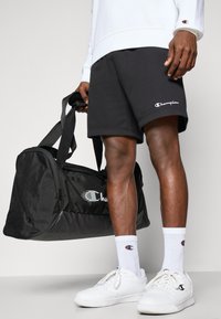 Schwarze Sporttasche mit Champion-Logo, getragen mit grauen Shorts und weißem Sweatshirt. Weiße Sneakers und gerippte weiße Socken mit Logo.