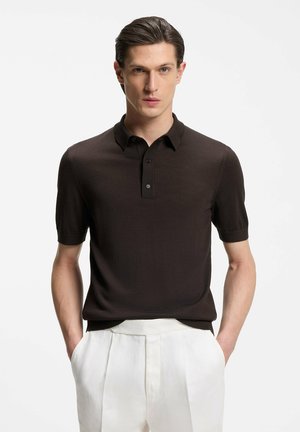 Uomo in piedi con le mani nelle tasche che indossa una polo a maniche corte marrone scuro e pantaloni bianchi plissettati su sfondo semplice.