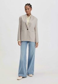 Beige maßgeschneiderter Blazer mit einem Knopf, zwei Fronttaschen, kombiniert mit hellblauen weitem Jeans und beigen Schuhen. Strukturiertes Stoffdetail.