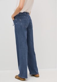 Široké deninové kalhoty v tmavě modré barvě s hladkým povrchem, s zadními kapsami a pasem na šňůrku. Nosí se s pohodlnými sandály.