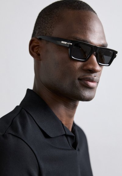 Ochelari de soare dreptunghiulari negri, cu rame groase, având un accent argintiu pe braț. Purtați cu un tricou polo negru, care prezintă o textură netedă.
