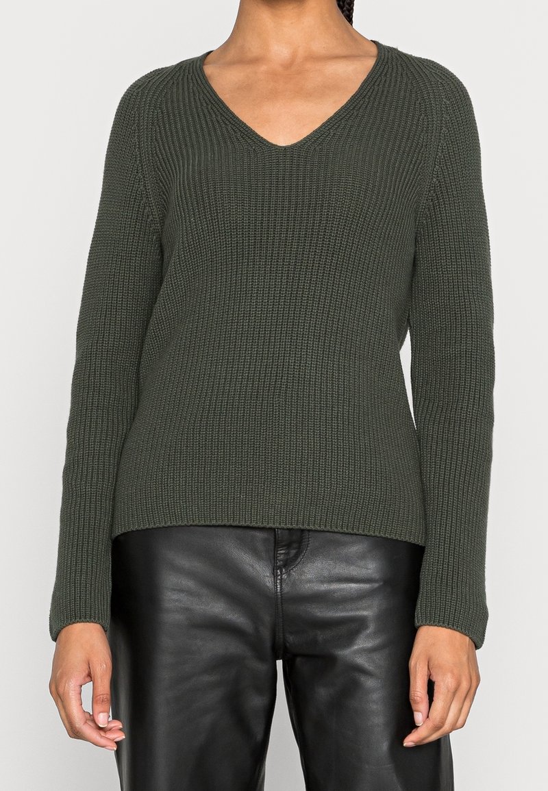 Nainen, joka pukeutunut tummanvihreään ribbineulottuun V-kaulaiseen neulepaitaan ja mustiin nahkahousuihin, kuvattuna kaulasta lantiolle yksinkertaisella taustalla.