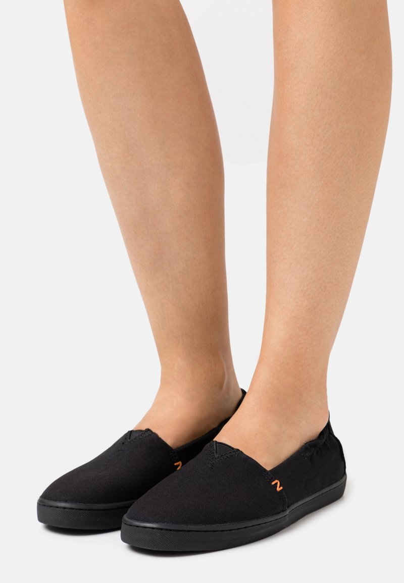 Fødder iført sorte slip-on lærredssko med gummisåler og lille orange logodetalje på siden.