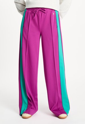 Persoon die wijde paarse broek draagt met turquoise zijstrepen en een elastische tailleband, gecombineerd met witte schoenen.