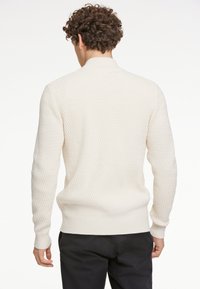 Ljusgräddfärgad texturerad stickad tröja med diamantmönster, långa ärmar och ribbad fåll, dragen över mörka byxor, bakifrån.