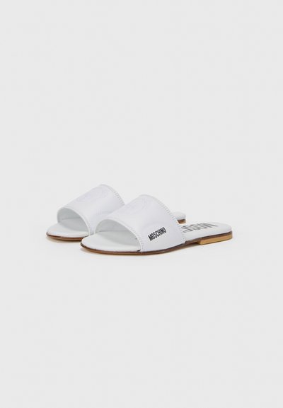 Sandales blanches à enfiler avec une tige en cuir présentant un logo en relief et des lettres noires "MOSCHINO" sur le côté, posées sur une semelle plate.
