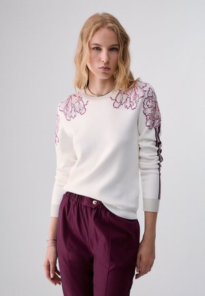 Giovane donna con capelli biondi ondulati che indossa un maglione bianco con ricamo floreale viola e pantaloni viola abbinati su uno sfondo semplice.