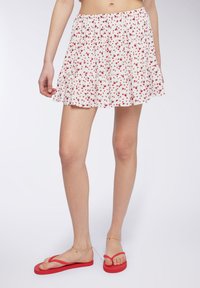Mini gonna bianca con motivo floreale rosso, volant sovrapposti e vita elastica. Abbinata a ciabatte rosse e catena alla caviglia.