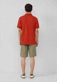 Rotes Poloshirt mit kurzen Ärmeln, kombiniert mit beigen Shorts. Das Model trägt lässige Schuhe mit weißen und dunklen Akzenten und steht vor einem grauen Hintergrund.