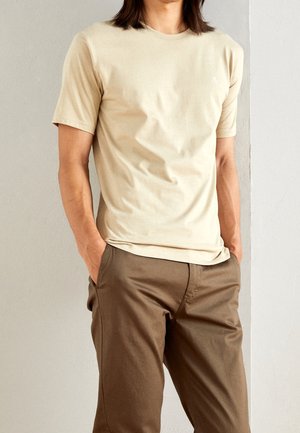 Beige cotton t-shirt with short sleeves and a crew neckline, worn with brown pants. The shirt features a small logo on the chest.