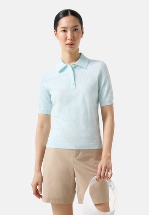 Kobieta ubrana w jasnoniebieską koszulkę polo z krótkim rękawem i beżowe szorty, trzyma białą daszkową czapkę w prawej ręce.